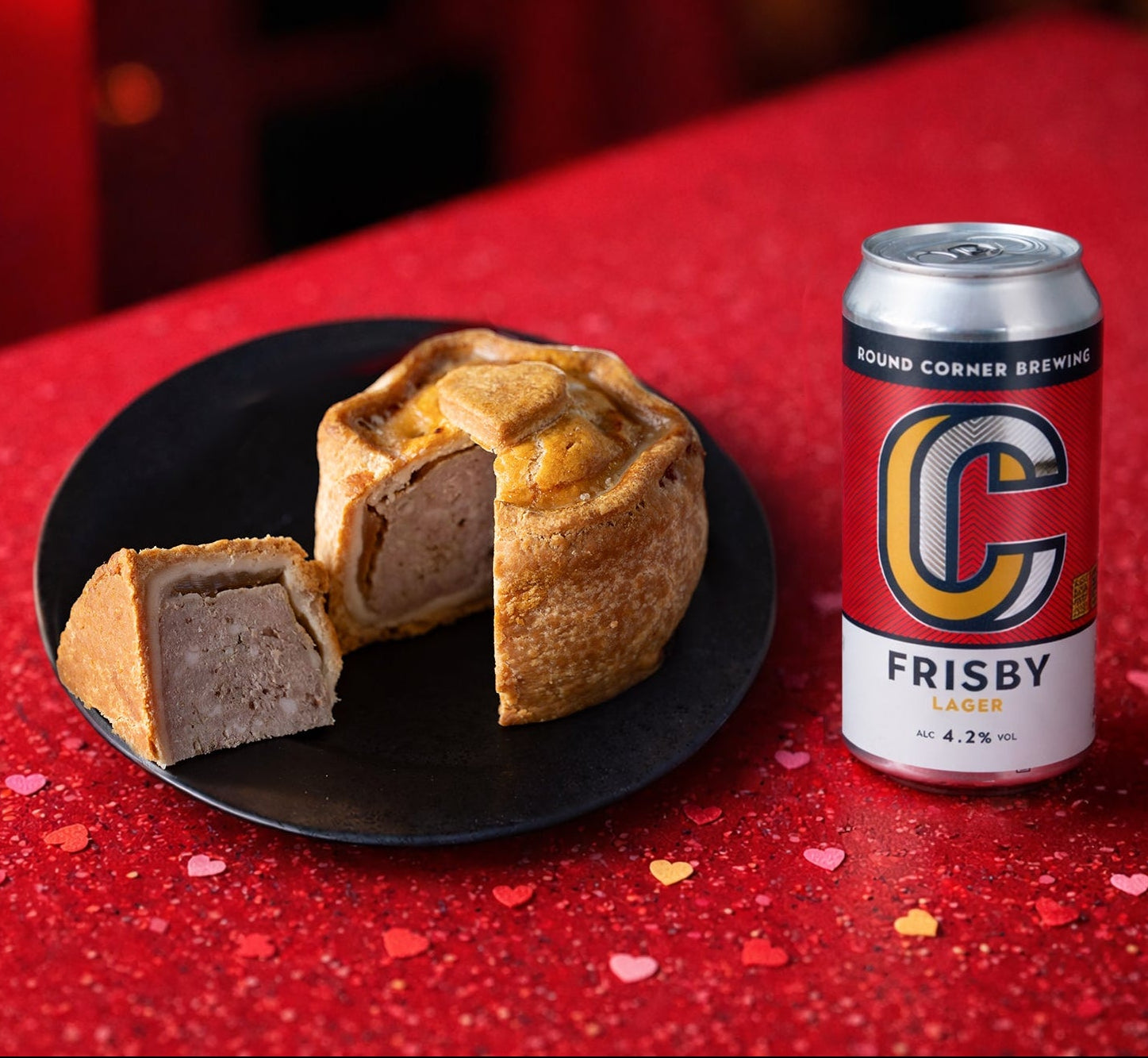 Love Heart pork pie with a slice removed on a black plate next to a can of Frisby beer on a red table cloth with hearts.